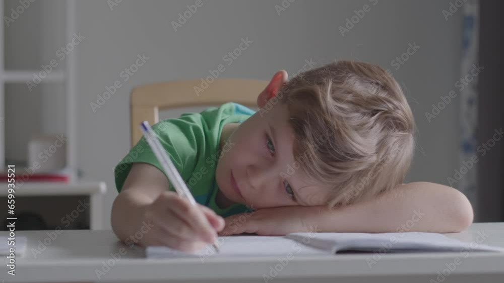 A bored tired sad upset little child boy kid son schoolboy schoolchild student at the table desk writing homework boring class lesson drawing