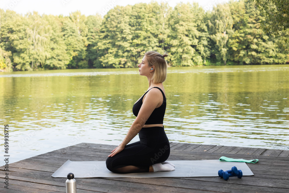 Woman doing exercises in nature near the lake. woman practicing yoga. The girl is engaged in fitness. The girl performs asanas. Yoga mat, bottled drinking water and stretch elastic band.