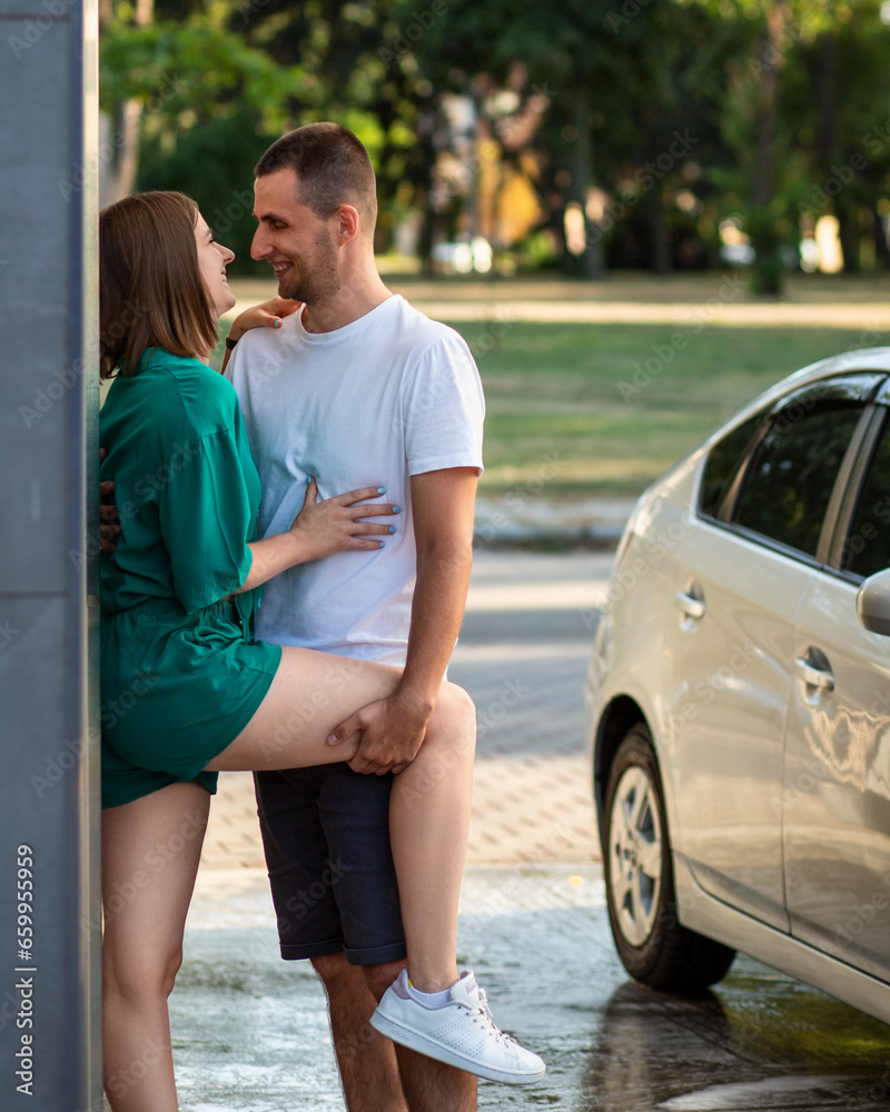 Young pretty caucasian couple playing hugging and kissing near a wall on an automatic car wash, laughing and having fun together