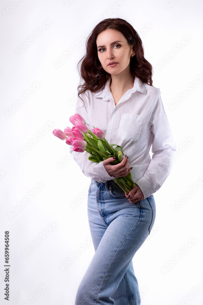 Studio portrait of a beautiful sexy slim young caucasian brunette in white shirt and blue jeans standing with a bouquet of pink tulips Studio portrait of a beautiful sexy slim young caucasian brunette in white shirt and blue jeans standing with a bouquet of pink tulips