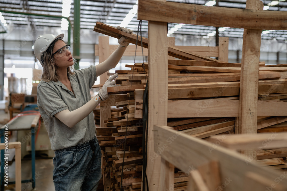 Confident female worker working in lumber warehouse of wooden furniture factory checking stock. Skilled carpenter inspector wears safety hardhat examining hardwood material for production facility. Confident female worker working in lumber warehouse of wooden furniture factory checking stock. Skilled carpenter inspector wears safety hardhat examining hardwood material for production facility.
