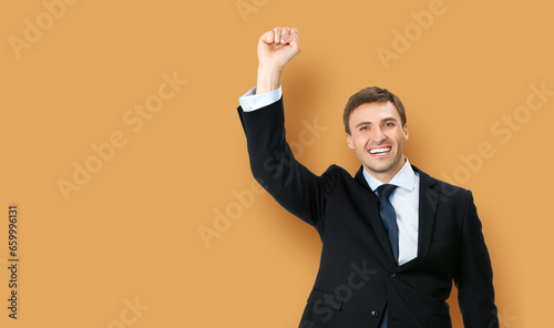 Image of very happy, excited gesturing business man in black confident formal suit, necktie, raising hand fist, on brown bricks wall office background. Advertisement ad concept photo