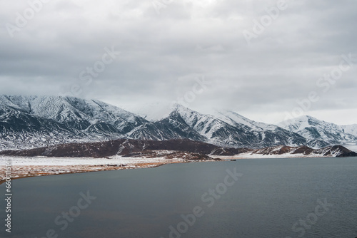 Snow-covered winter pastel colors landscape. View of the dark lake and snow hills. Cold winter weather. Harsh climate background in Mongolia.