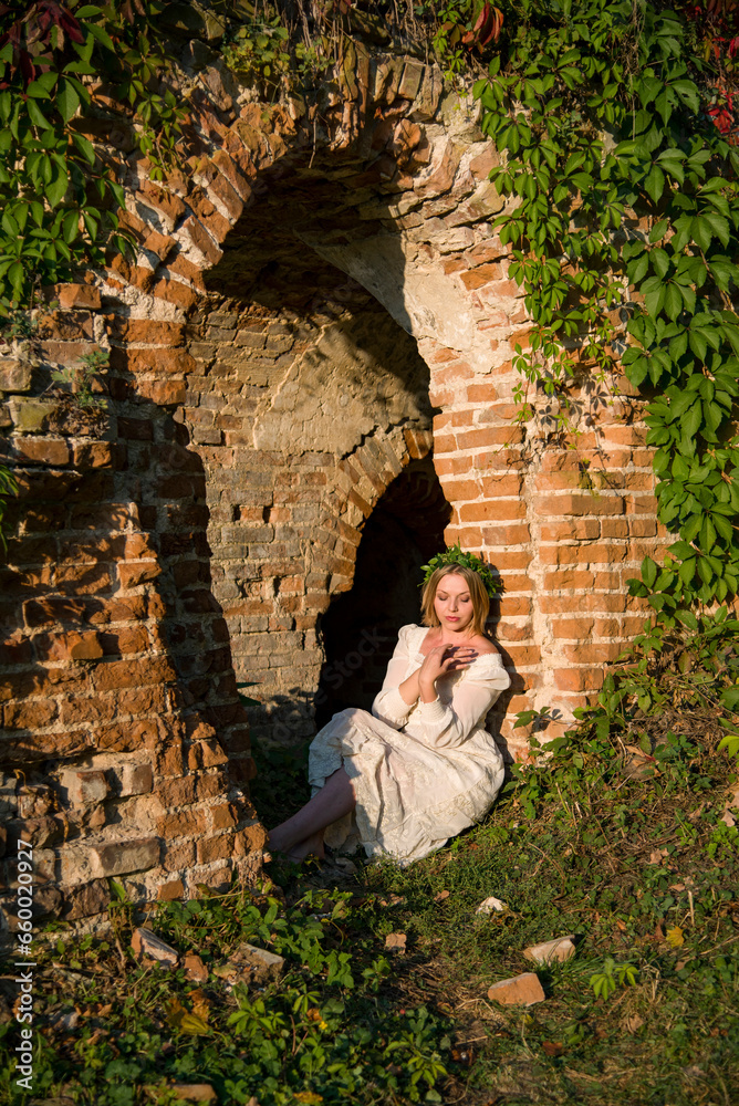 barefoot beautiful blonde woman in a white dress and wreath from green leaves sitting in ruins near destroyed arch among the weaving branches on sunny autumn evening