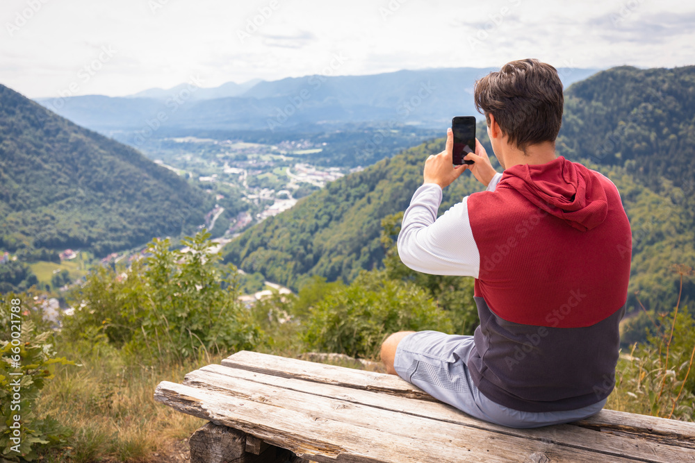 Hiking man sitting on a bench at a viewpoint enjoying taking photos of beautiful mountains and valley landscape with a smartphone