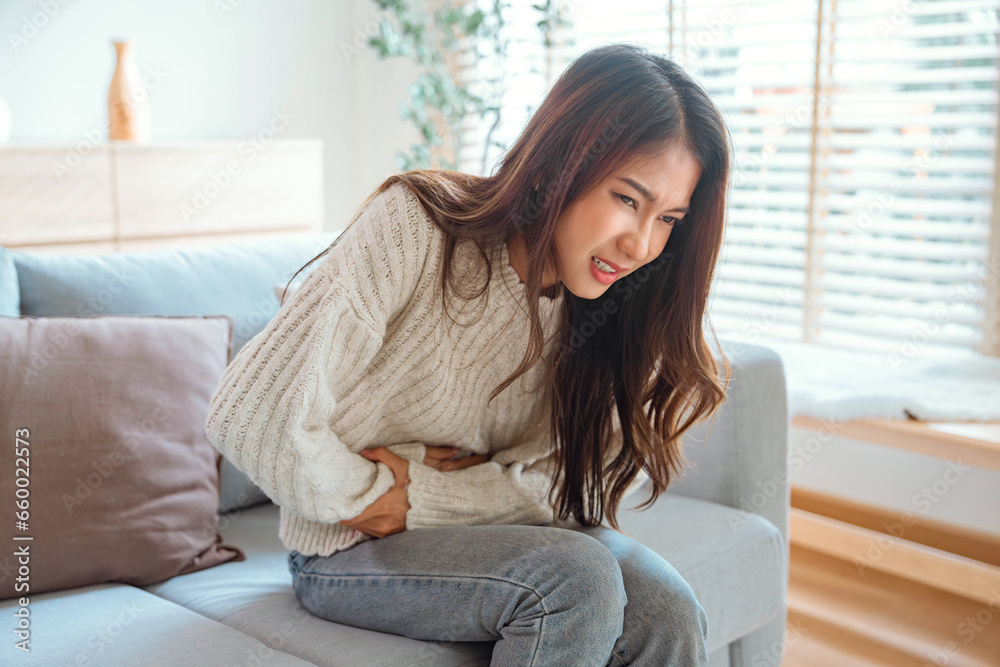 Sick young Asian woman suffering from stomach ache sitting on sofa in living room at home, Abdominal pain, gastritis, and painful periods concept. Sick young Asian woman suffering from stomach ache sitting on sofa in living room at home, Abdominal pain, gastritis, and painful periods concept.