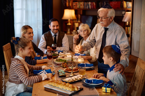 Jewish senior man lighting menorah during family meal at dining table on Hanukkah.