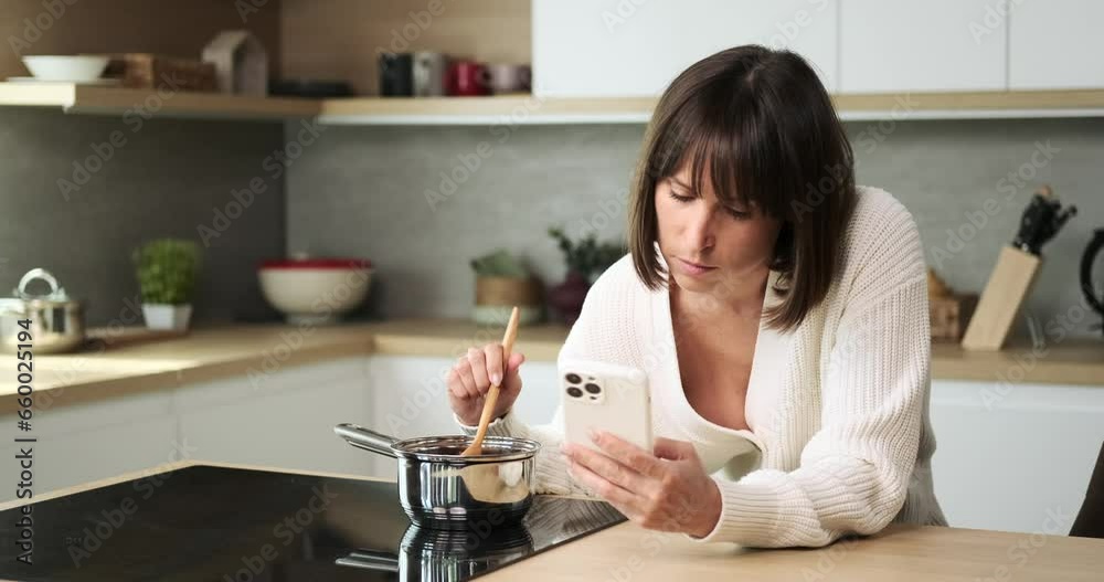 A bored Caucasian woman multitasks by cooking while casually browsing her phone. Her demeanor suggests a moment of leisure, combining culinary activities with digital entertainment.