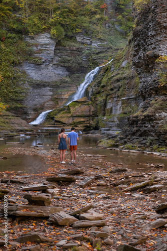 Young Asian couple walking to the waterfall in deep gorge surrounded by autumn forest with bright yellow and orange leaves. Long exposure, wide angle shot. Robert H Treman State Park, NY, USA.
