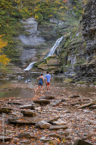Young Asian couple walking to the waterfall in deep gorge surrounded by autumn forest with bright yellow and orange leaves. Long exposure, wide angle shot. Robert H Treman State Park, NY, USA.