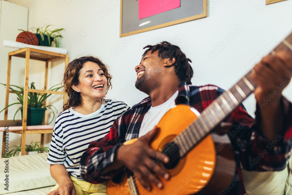 Fototapeta premium Happy young couple singing a song playing guitar on sofa in living room at home