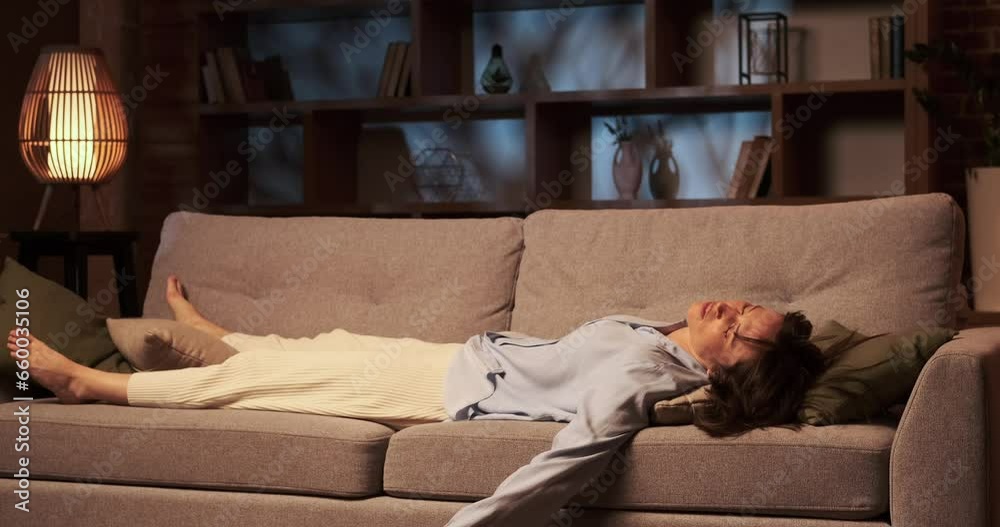 Weary Caucasian woman, drained from the day endeavors, collapses onto the sofa in the living room. Her face carries the weight of fatigue, and body surrenders to the softness of the couch.