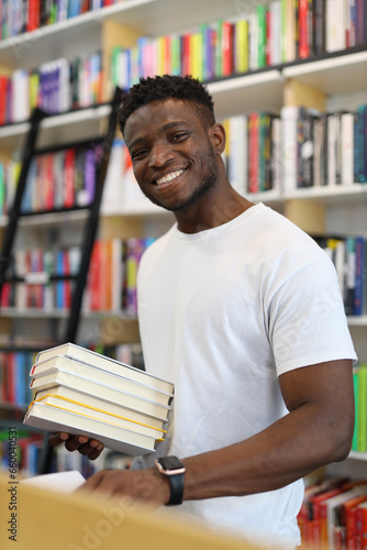 A cheerful young student in a university library, surrounded by books on bookshelves, stands confidently, studying with intelligence.