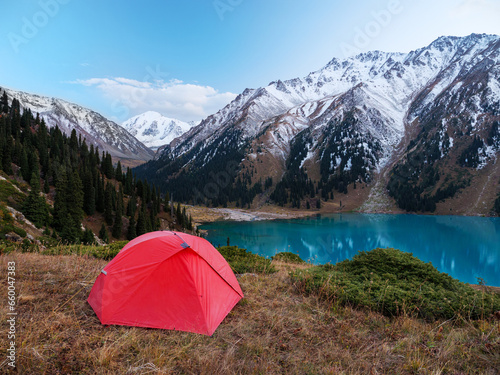 Camping with a red tent in the highlands on the shore of a beautiful lake. Panoramic views of snow-capped peaks and turquoise lake