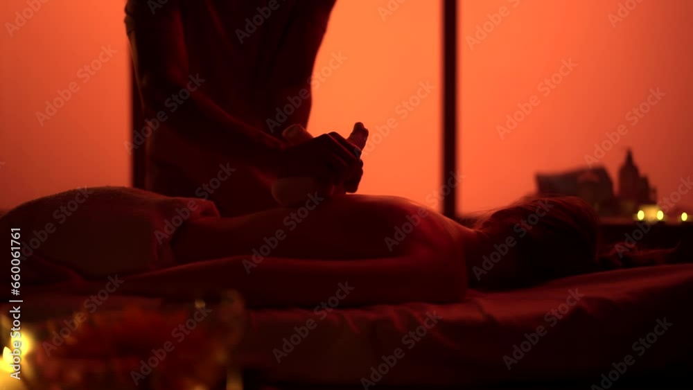 Masseur, massage specialist giving a massage to his patient using herbal bags. Silhouettes in the massaging room, spa procedure. Exotic, orange neon lights. Masseur, massage specialist giving a massage to his patient using herbal bags. Silhouettes in the massaging room, spa procedure. Exotic, orange neon lights.