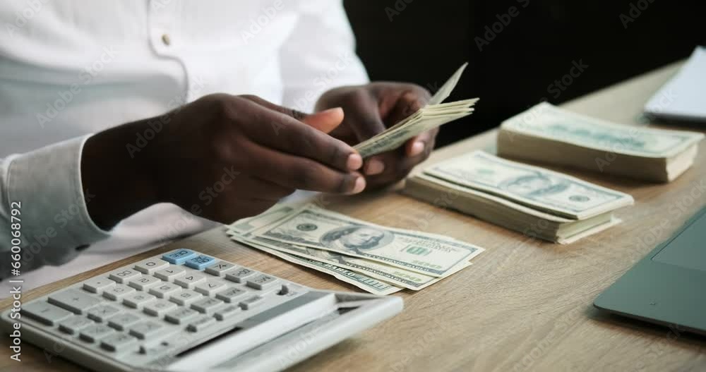 This close-up shot captures the hands of a focused Black man as he meticulously counts cash using a calculator. His careful and precise financial management is highlighted in this detailed view. This close-up shot captures the hands of a focused Black man as he meticulously counts cash using a calculator. His careful and precise financial management is highlighted in this detailed view.
