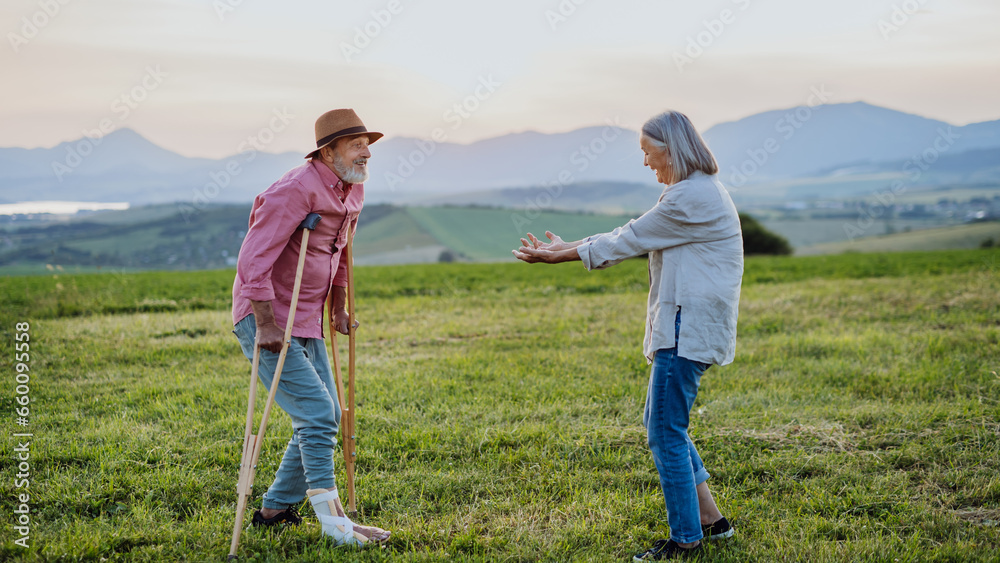 Senior man with broken leg and crutches. Recovery, rehabilitation after injury or surgery in the nature. Wife helping husband with walking during easy walk on their vacation.