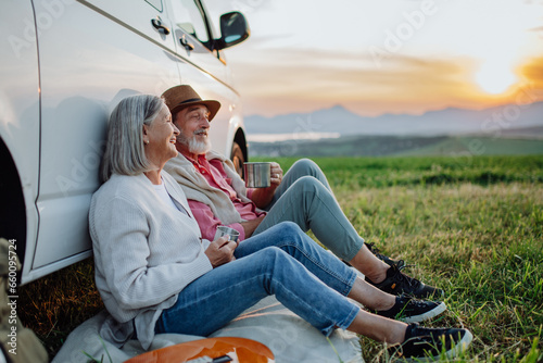 Senior couple sitting by car and drinking coffee after long drive during their roadtrip. Elderly spouses at autumn roadtrip watching the sun set behind High Tatras.