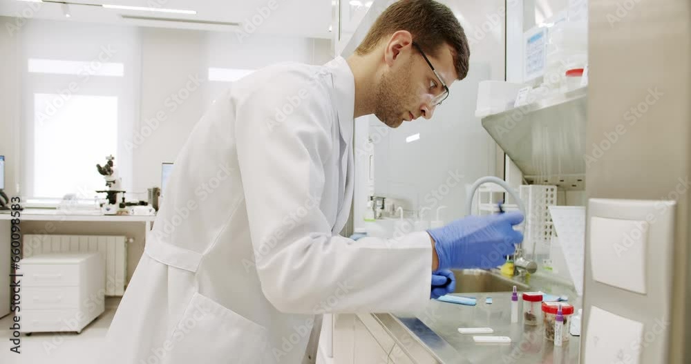 Modern Medical Research Laboratory. Handheld shot of man in white robe with gloves and glasses putting vial with sample into machine during work in medical laboratory in clinic. Modern Medical Research Laboratory. Handheld shot of man in white robe with gloves and glasses putting vial with sample into machine during work in medical laboratory in clinic.
