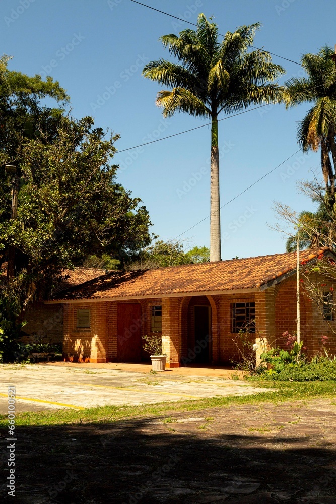 Casa com varanda, paredes de tijolo a vista, talhado de telha de barro e muitas árvores no jardim.