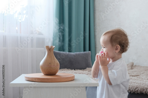 Water diffuser, wooden jug with steam. A girl of European appearance, two years old, joyful, in white clothes in the room. Blurred background. Concept of health care, air humidification