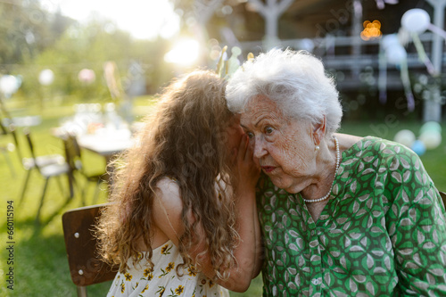 Girl whispering a secret into grandmother's ear at a garden party. Love and closeness between grandparent and grandchild. photo