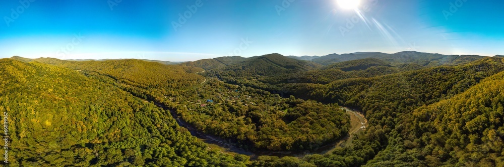 Fototapeta premium a farm near a river bend surrounded by the Western Caucasus mountains covered with green deciduous forest on a sunny day in early autumn - aerial panorama