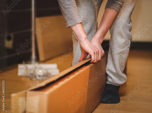 The hands of a caucasian young man in a gray tank top with sleeves open a large cardboard box