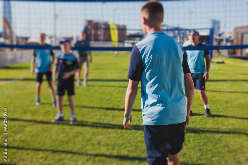Obraz premium Volleyball game, junior teenage school team of kids play volleyball, players on the outdoor playground with net and green lawn grass court, sports children team during the game, summer sunny