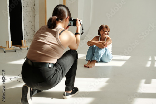 Happy African American girl in casualwear posing for camera while sitting on the floor by wall in front of photographer during photo session © pressmaster