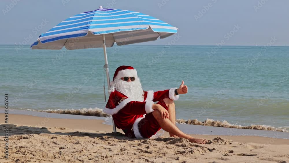 Santa Claus sits under a beach umbrella against the background of the sea and enjoys a hot summer day. Christmas or New Year holidays in hot countries. Santa's summer vacation.