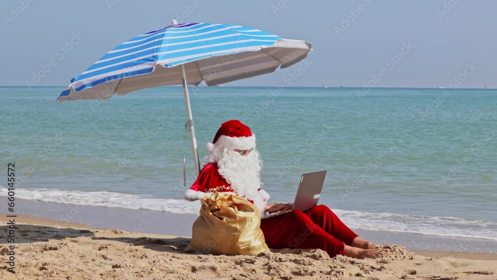 Santa Claus sits under a beach umbrella against the background of the sea on a hot summer day and types something on a laptop. Christmas or New Year holidays in hot countries. Santa's summer vacation. Santa Claus sits under a beach umbrella against the background of the sea on a hot summer day and types something on a laptop. Christmas or New Year holidays in hot countries. Santa's summer vacation.