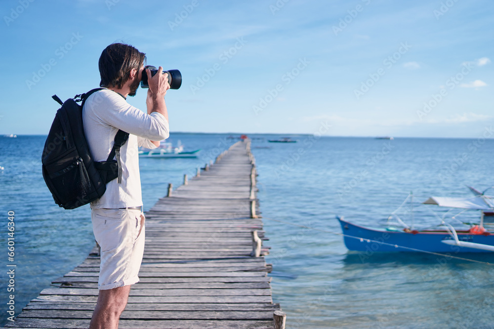 Obraz premium Photography and travel. Young man with rucksack standing on wooden fishing pier taking photo of beautiful tropical sea view.