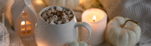 White mug with aromatic autumn hot drink - sweet delicious pumpkin latte with marshmallow and spices White background, bokeh lights, candles, knitted sweater as decor. Banner