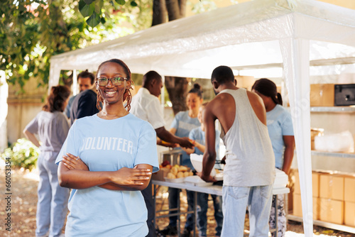 Young black woman with glasses stands outdoors, arms crossed, looking at the camera. Diverse group of volunteers supports a non-profit program dedicated to hunger relief and helping needy individuals. photo