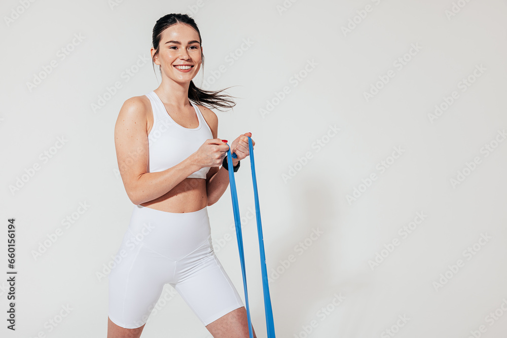 Cheerful female in white fitness attire exercising with blue resistance band at a white wall Cheerful female in white fitness attire exercising with blue resistance band at a white wall