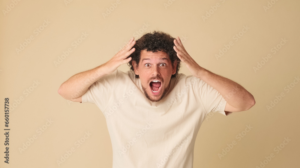 Shocked guy with curly hair dressed in beige t-shirt looking in surprise at the camera with big eyes, raises his hands to his face, wow, isolated on beige background in the studio Shocked guy with curly hair dressed in beige t-shirt looking in surprise at the camera with big eyes, raises his hands to his face, wow, isolated on beige background in the studio