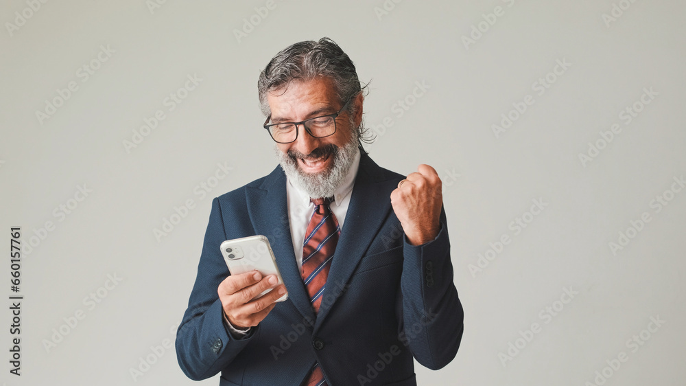 Businessman in glasses with gray hair, looking with smile at the mobile phone screen, getting good deal, winning, isolated on gray background in the studio Businessman in glasses with gray hair, looking with smile at the mobile phone screen, getting good deal, winning, isolated on gray background in the studio