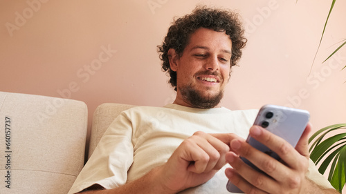 Close up, positive young man with curly hair and beard, sitting on sofa in cozy living room, scrolls his mobile phone on the screen