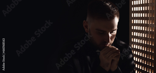 Portrait of young praying priest in confession booth