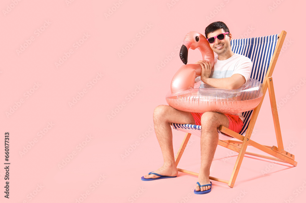 Fototapeta premium Young man with swim ring sitting in deck chair on pink background