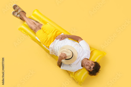 Young man with hat and swim mattress relaxing on yellow background, top view