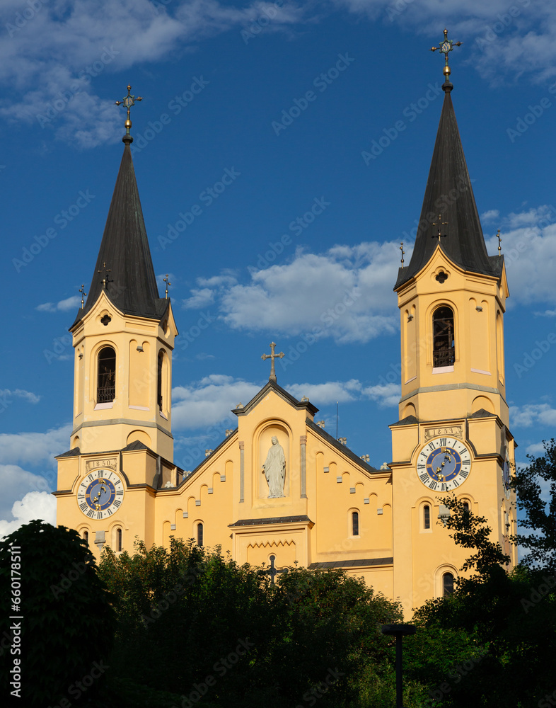 View of Church of Assumption of Mary with statue of Virgin Mary in niche on main facade of Romanesque building and two steeples rising above green trees against blue sky in Italian town of Bruneck