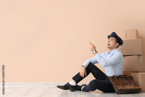 Young postman with bag and parcels pointing at something near beige wall