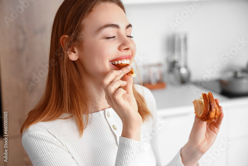 Beautiful young woman eating tasty croissant in kitchen