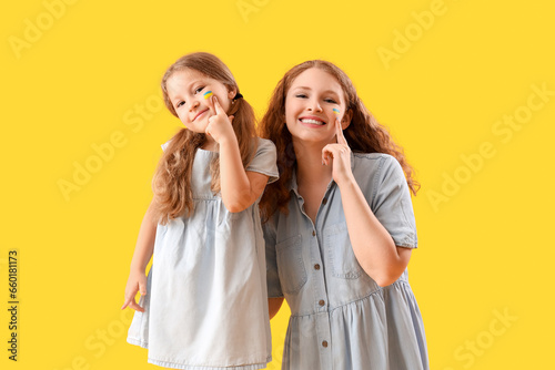 Happy little girl and her mother pointing at drawn Ukrainian flags on their faces against yellow background