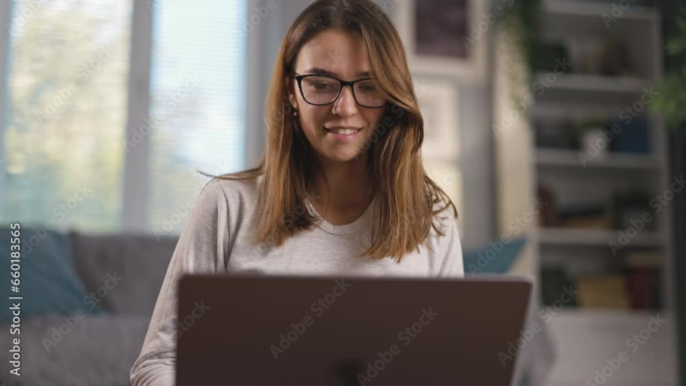 Pretty young woman sits on carpet, put laptop on laps, browses and watches captivating material, smiling with relaxed demeanor