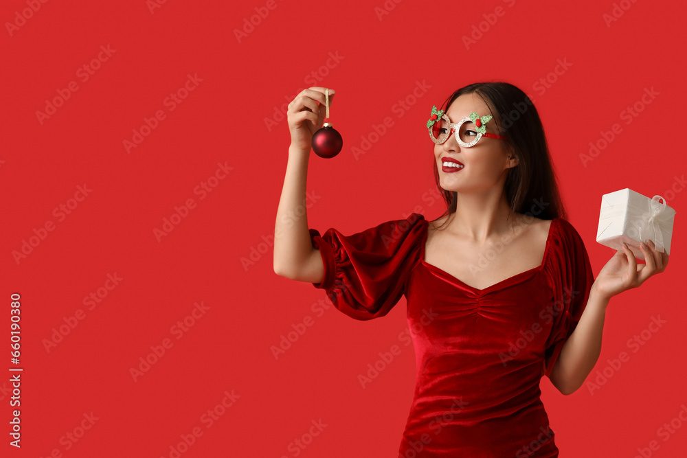 Beautiful young Asian woman in party glasses with Christmas ball and gift box on red background