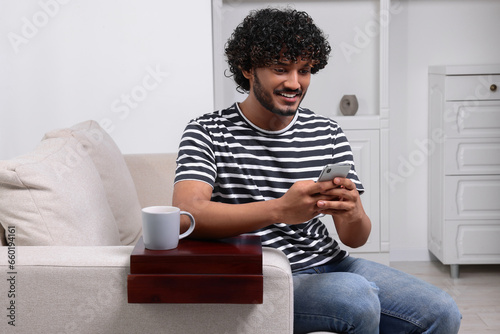 Happy man using smartphone on sofa with wooden armrest table at home