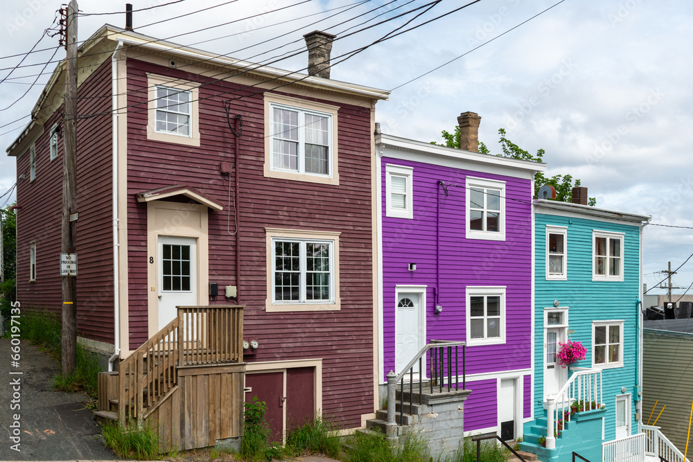 Street view of multiple colorful wooden residential buildings of various colors. The historic houses are row or adjoined. The buildings are on a hill with a narrow sidewalk in front of the old homes.